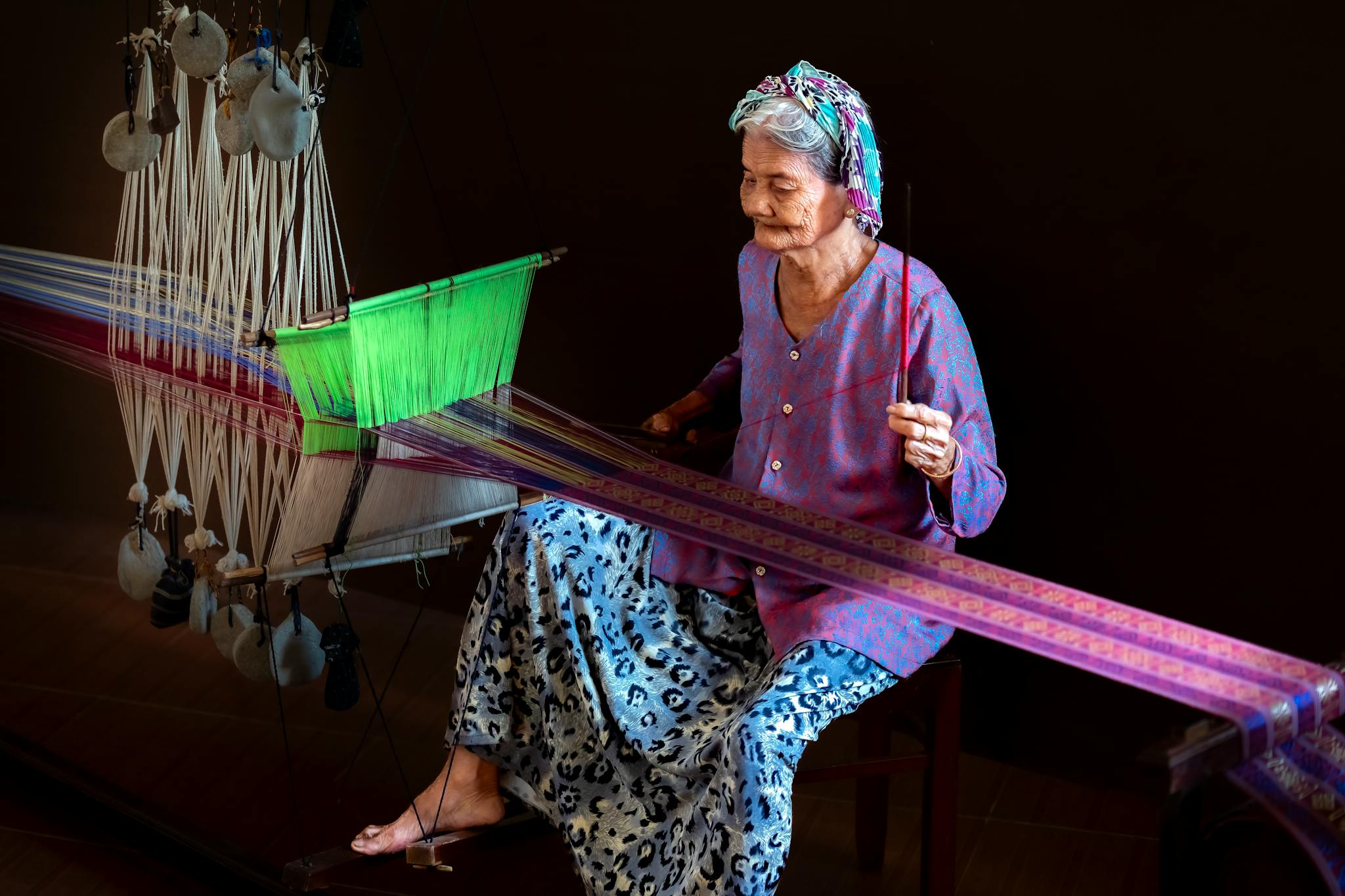 Elderly woman skillfully weaving vibrant fabric on a traditional loom indoors.